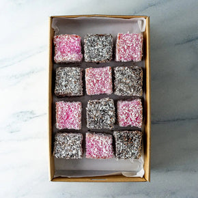 A rectangular cardboard box of 12 mini lamingtons in chocolate and raspberry flavours, arranged in a grid pattern, on a marble table.