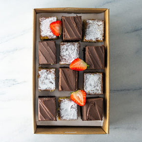 A rectangular cardboard box filled with a mix of 12 sweet slices in orange and almond, and chocolate mud flavours, garnished with fresh strawberry on a marble table.