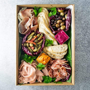 A large rectangular cardboard box containing antipasto, including salami, prosciutto, mortadella, olives, dips, pickled vegetables, and lavosh, on a marble table.