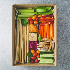 A box containing three dips, batons of celery, carrot, cucumber, red and green pepper, cherry tomatoes, crostini, and grissini sticks.