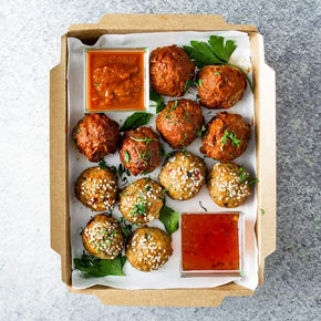 A rectangular cardboard box containing 12 mini meatballs including flavours like Thai chicken and Sicilian beef and pork, with tubs of dipping sauces, and parsley garnish, on a marble table.