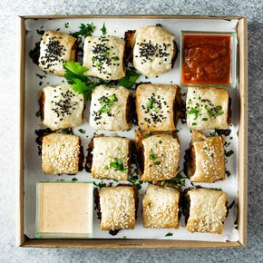 A square cardboard box filled with 12 mini sausage rolls with fillings like pork, chicken, beef, or sweet potato and lentil, along with tubs of dipping sauces on a marble table.