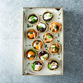 A rectangular cardboard box with a mix of mini savoury tarts including flavours like hot smoked salmon, beetroot, cherry tomato and caramelised onion, on a marble table.