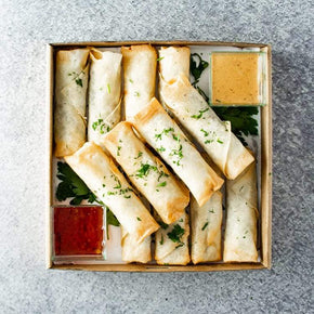 A square cardboard box containing 12 golden spring rolls including flavours like peking duck, barbecue pork, and vegetable, with tubs of dipping sauces, and parsley garnish on a marble table.
