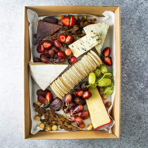 A large rectangular cardboard box containing wedges of 
Australian and European cheeses, quince paste, fresh & dried fruits, and crackers on a marble table.
