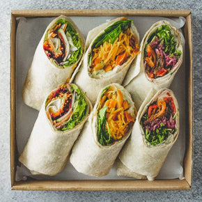 A square cardboard box containing three vegan savoury wraps cut in half on a marble table. Wraps include fillings like pumpkin and rocket, eggplant and caramelised onion, and avocado and tomato relish.