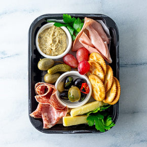 A rectangular container with an individual serve of antipasto and cheese, including slices of salami, mortadellam, and cheese, dip, crackers, olives, cornichons, and caper berries, on a marble table.