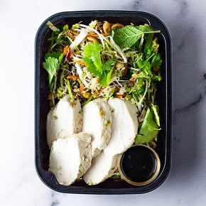 A small rectangular container with slices of lime poached chicken, snow pea, mung bean, and rice vermicelli salad with lime soy dressing on a marble table.