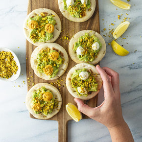 A person's hand reaching for a round pita bread topped with smashed avocado, dukkah, and whipped goats cheese on a wooden board, with lemon wedges scattered around.