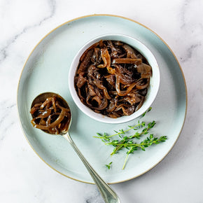 A top-down view of Balsamic Caramelised Onions in a white bowl and on a spoon, served on a light blue plate with fresh thyme on a white marble surface.