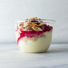 A clear plastic tub filled with bircher muesli topped with yogurt, berry compote, and toasted coconut.