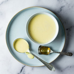A top-down view of a light blue plate on a white marble surface, holding a white bowl filled with Brandy Anglaise, alongside two spoons.