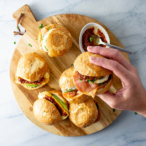 Six mini brioche breakfast sliders on a round wooden board, with a hand reaching for one of the sliders. The sliders are topped with various combinations of bacon and egg; salmon and egg; and avocado and  halloumi.