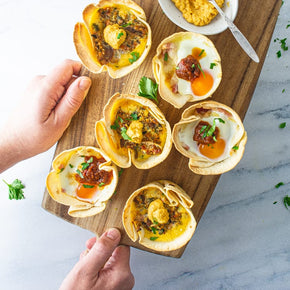 A person's hand holding a wooden board with breakfast pita cups with flavours like bacon, cheddar and cherry tomato with tomato relish; caramelised onion, sun dried tomato and ricotta with pumpkin hummus.