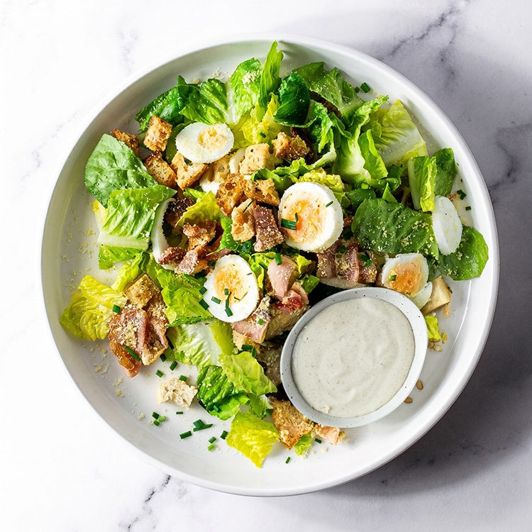 Plated salad with eggs, bacon, and croutons with dressing on a marble surface