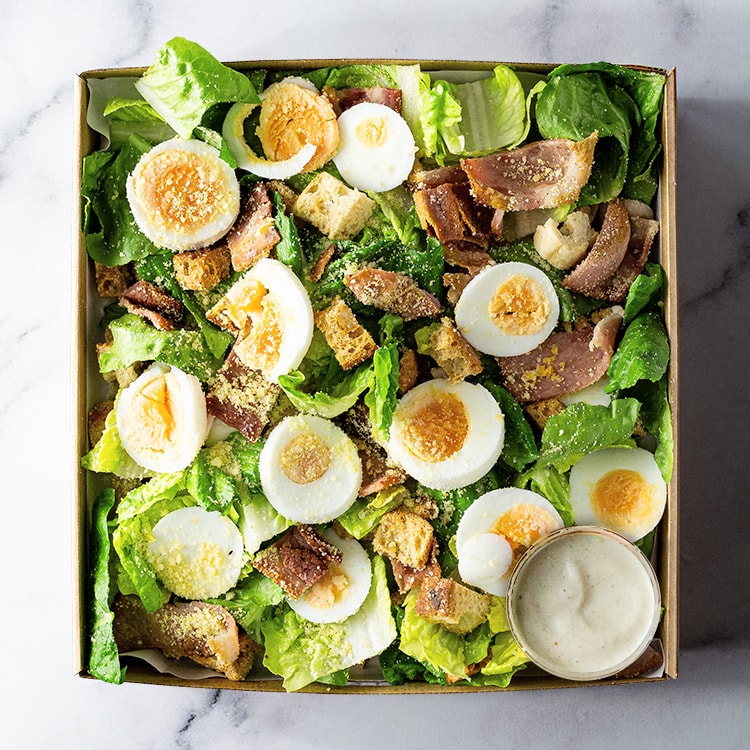 Box of salad with eggs, bacon, cos, croutons and dressing on a marble surface
