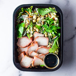 A small rectangular container with sliced char siu pork, wombok, Asian vermicelli slaw, and lime soy dressing on a marble table.