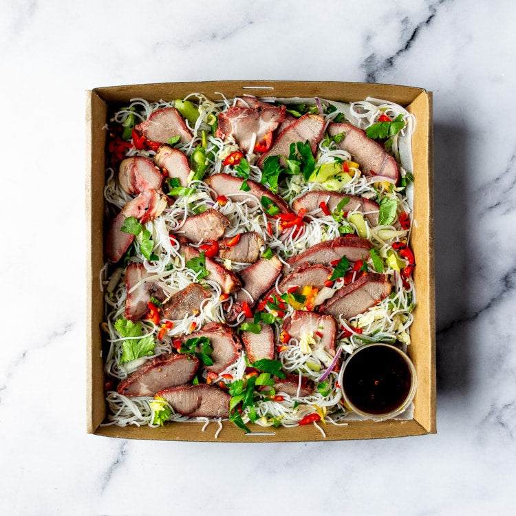 Box of char siu pork, rice vermicelli, capsicum, bean sprouts, wombok, celery, coriander, mint, and nigella seeds with a bowl of Nam Jim dressing on a marble background