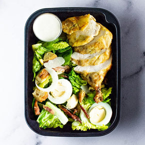 A smallrectangular container with slices of poached chicken, caesar salad and parmesan aioli dressing on a marble table.