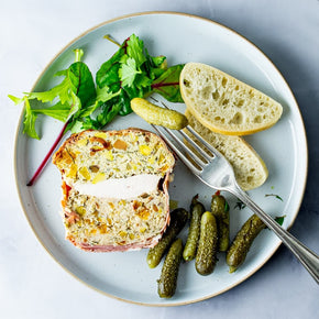 A plated food scene showing a slice of Chicken, Apricot & Pistachio Terrine with cornichons and bread slices on a light blue plate with a fork.