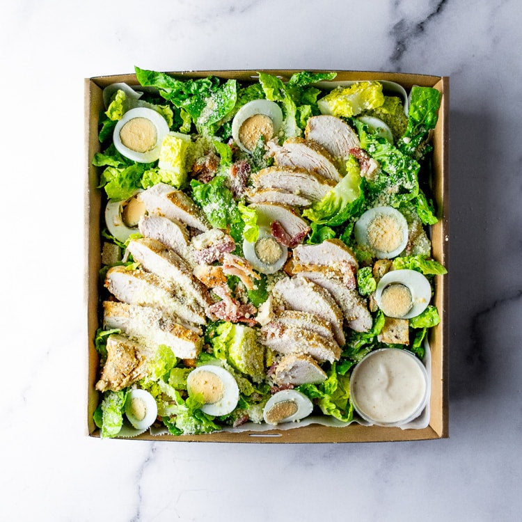 Box of Moroccan spiced chicken, cos, bacon, eggs, crispy croutons, and bowl creamy parmesan aioli on a marble background