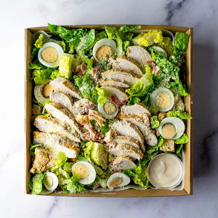 Box of salad with Moroccan chicken, cos, bacon, eggs, croutons, and parmesan on a marble table