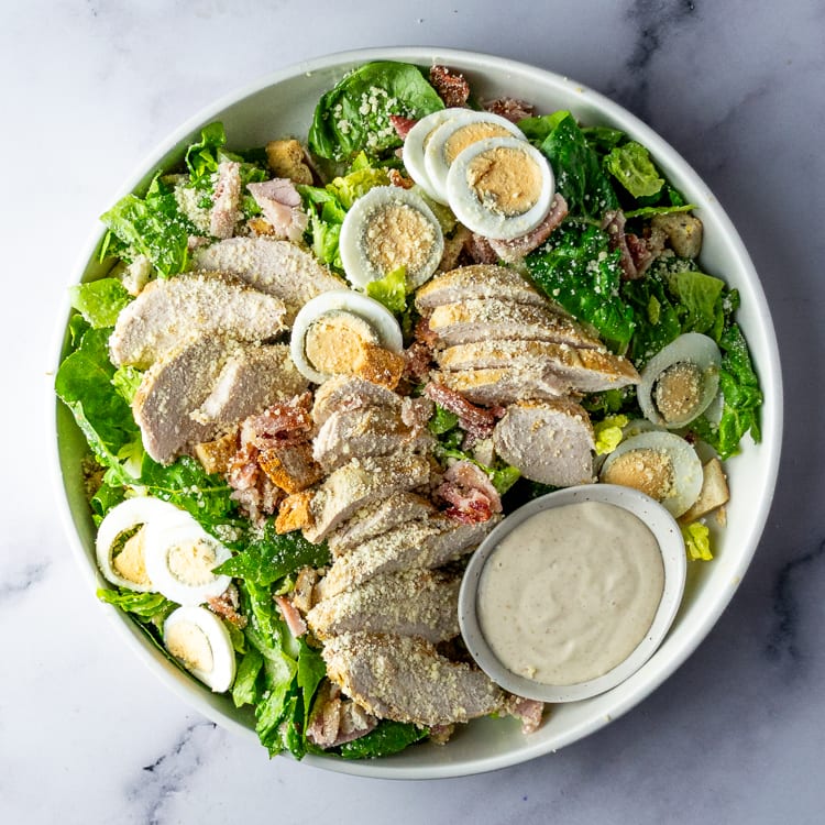 Moroccan chicken, cos, bacon, eggs, croutons, and parmesan aioli in a large salad bowl on a marble table