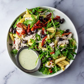 Shredded chicken, penne, sun-dried tomatoes, kalamata olives, peppery rocket, and pesto mayonnaise in a large salad bowl on a marble table