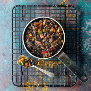 A top-down view of a Christmas Fruit Mince in a small metal pan with a long handle, resting on a black wire cooling rack over a textured teal surface. A silver teaspoon placed beside the pan.
