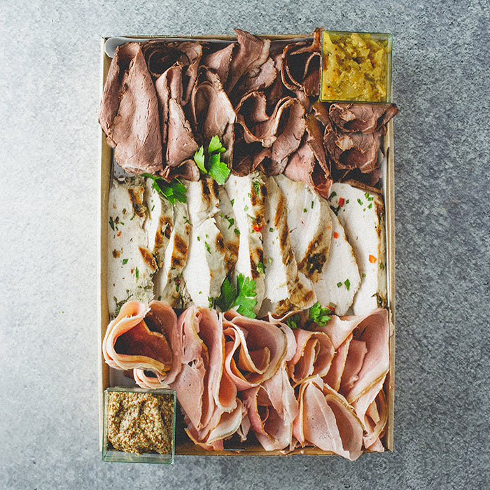 A rectangular cardboard box containing slices of rare roast beef, poached chicken breast and leg ham, along with baguette and tubs of mustard and chutney on a marble table.