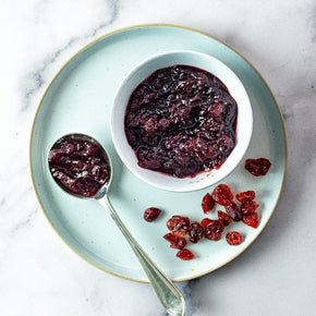 A top-down view of Cranberry & Orange Sauce in a small white bowl, with a spoonful beside it and scattered dried cranberries on a pale blue ceramic plate, set on a light marble surface.