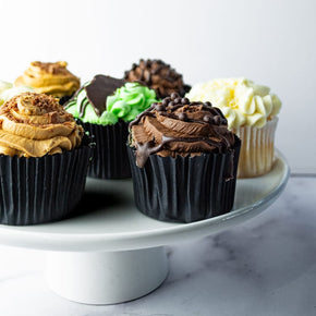Side view of a white cake stand with eight decorated cupcakes in flavours like chocolate, caramel, mint, and vanilla on a white cake stand on a marble table with a light grey background.
