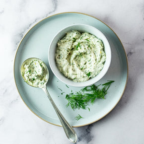 A top-down view of a Dill & Caper Cream in a white bowl on a light blue plate, with a spoonful of the cream and fresh dill on a white marble surface.