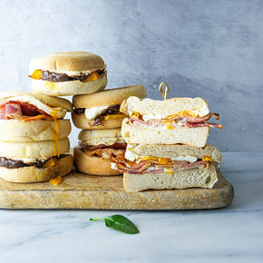 Side view of stacks of English muffins on a wooden board with fillings like bacon, egg and aioli; and pumpkin, onion, and goat's cheese, on a marble table.