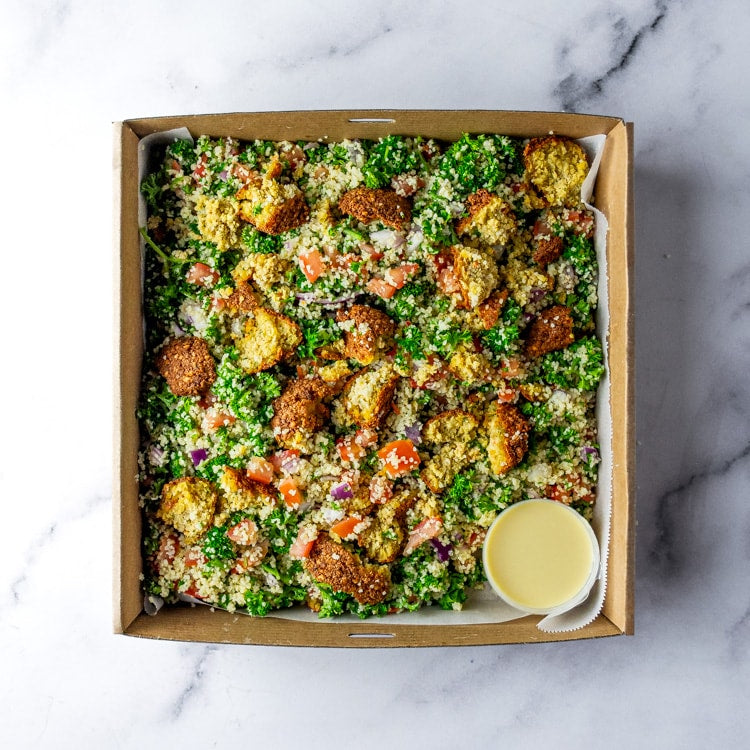 Box of falafel, quinoa, tomatoes, red onion, fresh parsley, and a bowl of lemon vinaigrette on a marble background