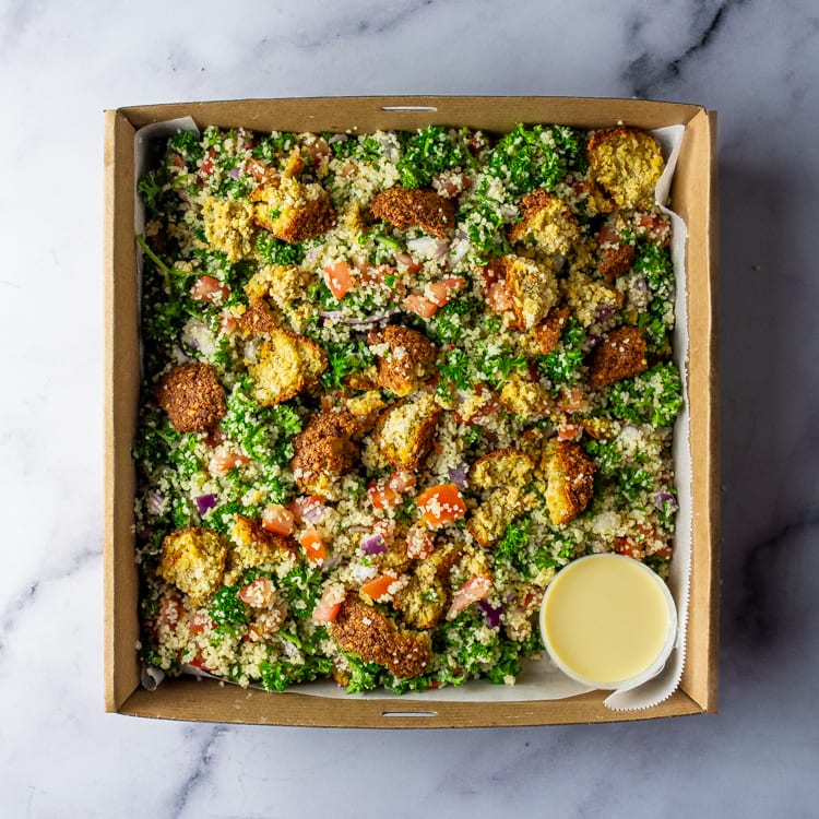 Box of salad with falafel, quinoa, tomatoes, red onion, fresh parsley, and lemon vinaigrette on a marble table