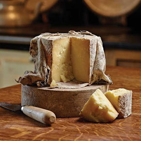 A rustic wheel of Ford Farm Cloth Bound Cave Aged Cheddar with a cut face showing its pale yellow interior resting on a wooden block with a knife on a wooden table.