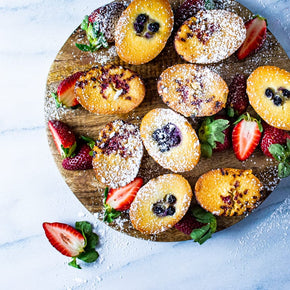 Friands in blueberry and raspberry flavours, garnished with powdered sugar and strawberries on a wooden board.