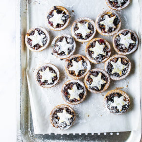 A top-down view of multiple Fruit Mince Pies on a baking tray lined with parchment paper. The pies have golden-brown pastry crusts, dark fruit filling, and a white star-shaped top dusted with powdered sugar.