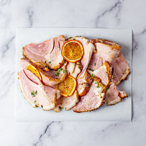 Sliced glazed ham arranged on a white rectangular board, garnished with thyme sprigs and dried blood orange slices, set on a light marble surface.
