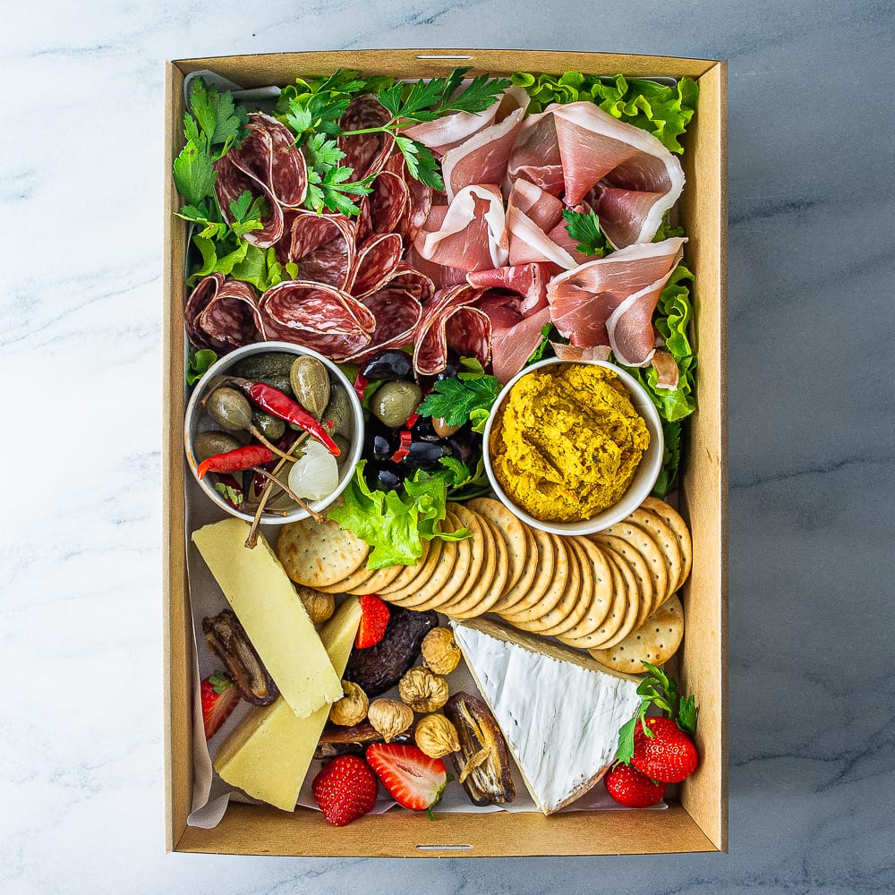 A rectangular cardboard box filled with slices of salami, prosciutto, and cheese, with tubs of dip, olives and pickled vegetables, along with crackers, fresh and dried fruit on a marble table.