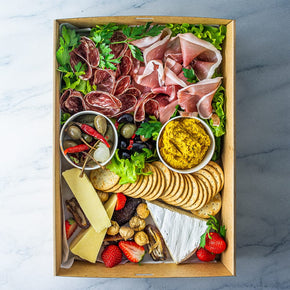 A rectangular cardboard box filled with slices of salami, prosciutto, and cheese, with tubs of dip, olives and pickled vegetables, along with crackers, fresh and dried fruit on a marble table.