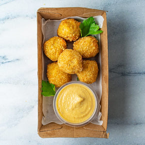 A small rectangular cardboard box containing six pumpkin arancini with a tub of cashew aioli, on a marble table.