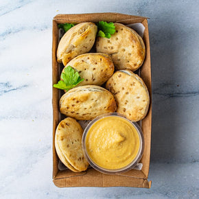 A small rectangular cardboard box containing six mini pies, a tub of cashew aioli, and parsley garnish, on a marble table.