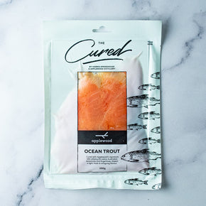 A 100g package of Harris Smokehouse Applewood Cured Ocean Trout, displayed on a light gray marble surface. The packaging is white with black text and illustrations of fish, featuring a clear window showing the orange-pink cured salmon inside.