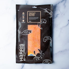 A 350g package of Harris Smokehouse Premium Smoked Salmon on a white marble background. The packaging is black with white illustrations of fish, a crab, and a shrimp, and features a clear window showing the orange-pink salmon inside.