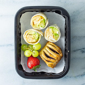 A black disposable container with three mini breakfast wraps, danish and fresh fruit on a marble surface.