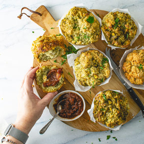 A persons hand reaching for one of six large savoury muffins along with a bowl of relish, teaspoon, and knife on a wooden board.
