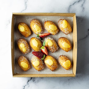 A square cardboard box of 12 lemon curd filled madeleines with strawberry garnish on a marble table.