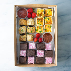 A rectangular cardboard box of mini quiche, tomato relish, sausage rolls, tomato sauce, sweet slice, mini lamingtons and fresh strawberries on a marble table.